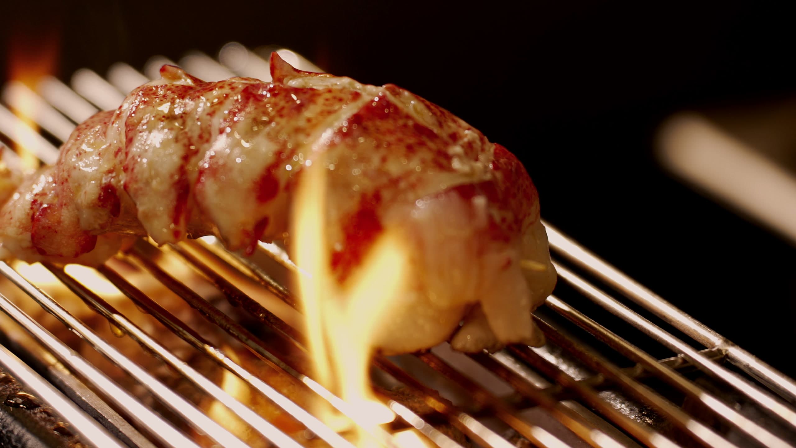 A glazed lobster tail on a skewer over a grill, flame rising through the grate beneath it