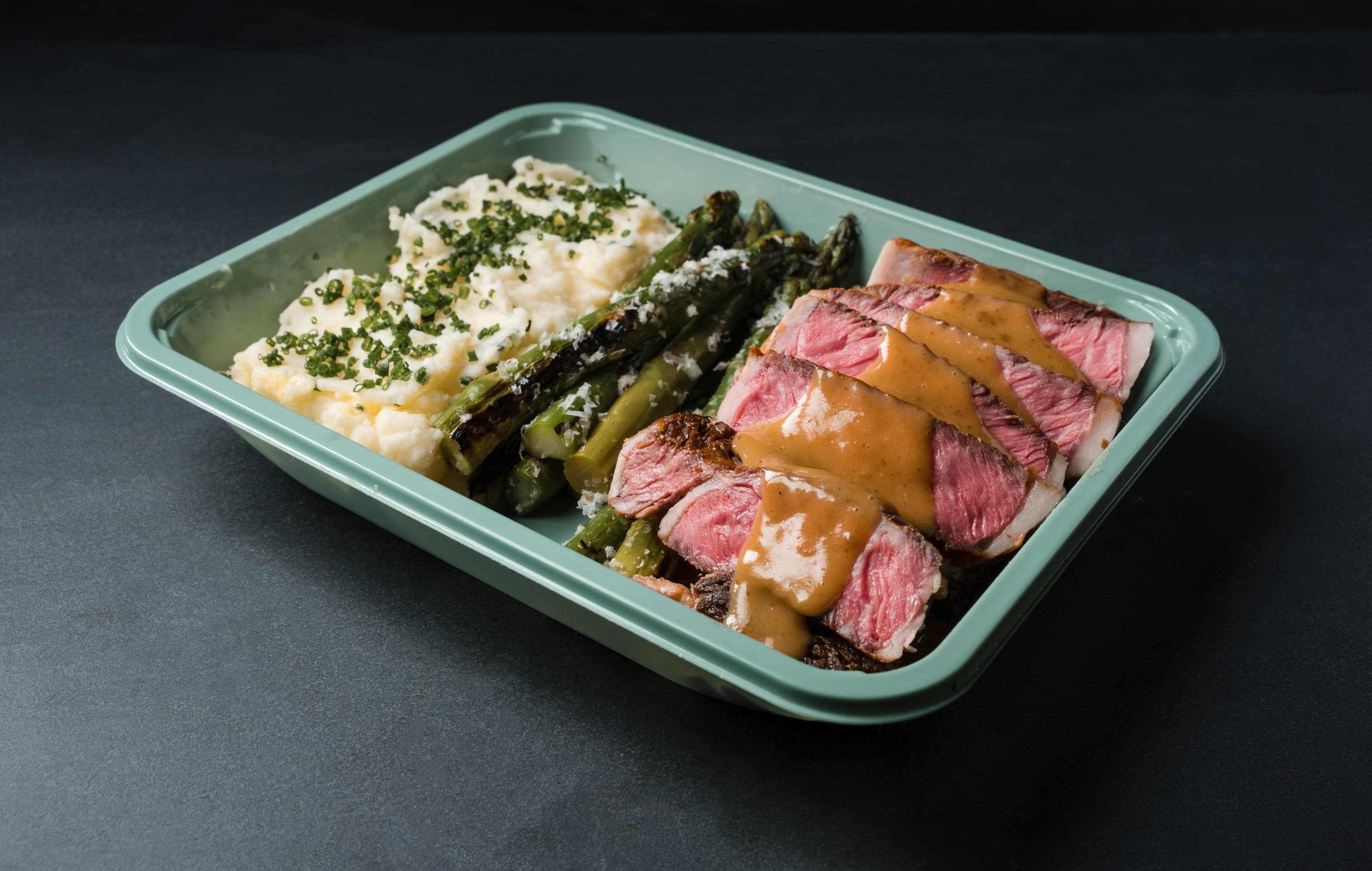 A to-go tray of sliced medium-rare strip steak with jus, grilled asparagus under parmesan and chive whipped potatoes